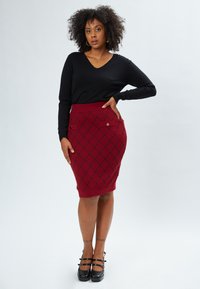Maglione nero a scollo a V abbinato a una gonna rossa aderente con un motivo a rombi e due tasche frontali. Scarpe con zeppa nera completano il look.