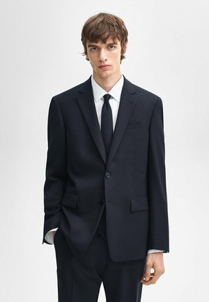 Dark navy suit with a tailored fit, single button, notch lapels, and two side pockets. Paired with a white shirt and black tie.