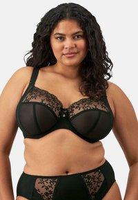 Sujetador negro con aros, top de encaje transparente y bordado floral, con tirantes ajustables y cierre central con gancho. Se muestran las bragas a juego.