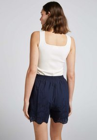 Débardeur blanc à bords festonnés associé à un short bleu marine avec broderies florales et un ourlet festonné. Ceinture élastique visible.