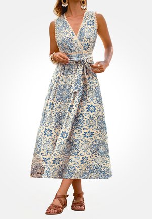Vestido crema sin mangas con estampado floral azul, escote en V, cintura anudada, llevado con pendientes grandes de concha, pulseras, anillos y sandalias trenzadas marrones.