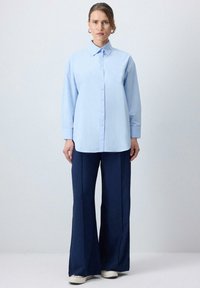 Camicia azzurra chiaro, con bottoni e maniche lunghe, colletto, abbinata a pantaloni larghi blu scuro e sneakers bianche.
