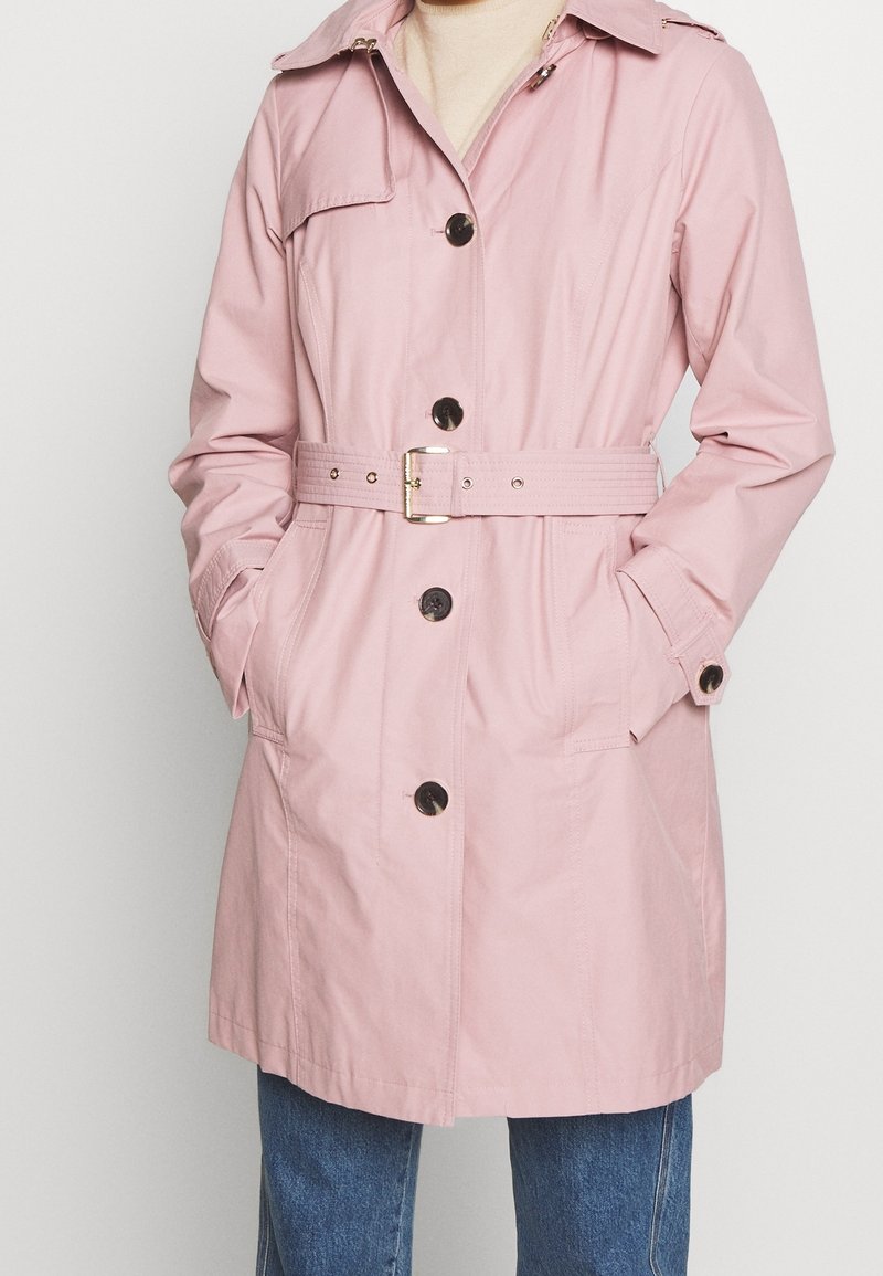 Personne portant un trench-coat rose clair ceinturé avec des boutons noirs et un jean bleu, debout les mains dans les poches.