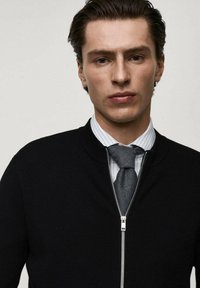 Crna zip-up pletena jakna s rebrastim ovratnikom, nosi se preko svijetloplavog prugastog košulje s dugim rukavima i sivoj kravati, prikazujući elegantan dizajn.