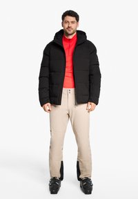 Veste matelassée noire avec capuche, couche intermédiaire rouge, pantalon de ski beige avec accents noirs, et chaussures de ski noires. Design simple, matériaux fonctionnels.