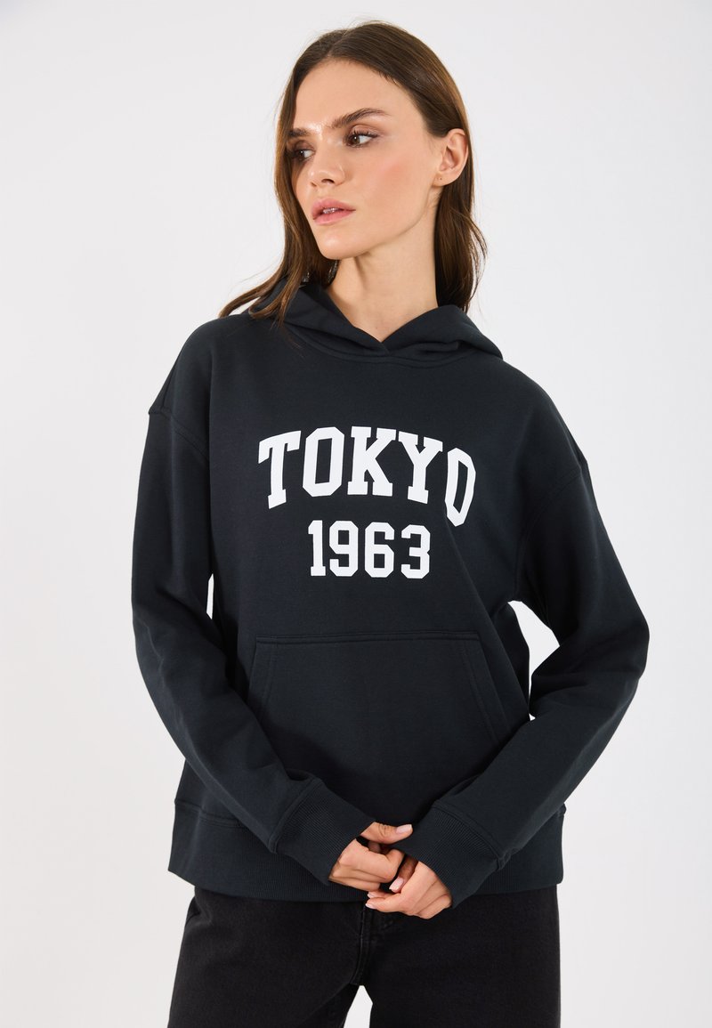 Zwarte hoodie met een buidelzak, voorzien van witte tekst "TOKYO 1963" over de borst. Het materiaal lijkt zacht met een ontspannen pasvorm.