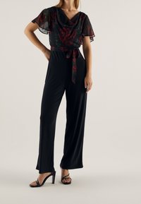 Combinaison noire avec superposition en tulle floral, manches courtes volantées, décolleté en V, taille ceinturée, jambes larges, et talons hauts noirs à lanières.