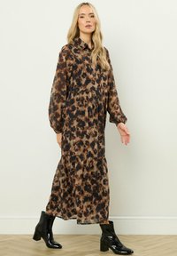 Blondynka w długiej sukience w brązowo-czarny wzór z długimi rękawami i czarnych botkach na kostkę, idąca po drewnianej podłodze.