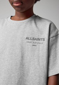 Hallitus hallist vahtidest valmistatud oversize t-särk, millel on must "ALLSAINTS" logo ja lisatekst jaapanikeeles, asudes vasakul rinnapiiril.