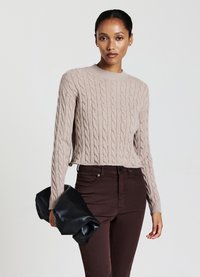 Pull en tricot beige avec col rond, porté avec un pantalon ajusté marron foncé ; tenant un sac à main en cuir noir. Texture lisse, accents côtelés.