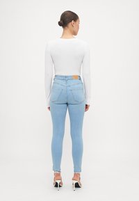 Jeans subțiri, de culoare albastru deschis, cu talie înaltă, buzunare la spate și o etichetă din piele maro la brâu. Poartați cu un top alb, cu mânecă lungă, ajustat.