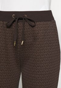 Bruna byxor med ett upprepat "MK" logomönster och svart dragsko knuten i en båge vid den ribbade midjebandet med guldfärgade ändar.