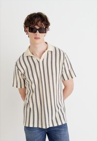 Polo a maniche corte color crema con strisce verticali blu navy, dotato di colletto a ribalta e tessuto texturizzato. Indossato con jeans scuri.