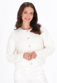 Cardigan alb scurt, realizat dintr-un material tricotat moale, având un guler rotund, închidere cu nasturi și tiv ondulat. Textură netedă cu o căldură subtilă.