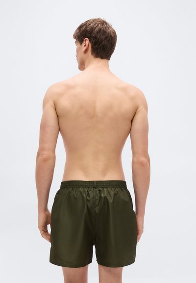 Νεαρός άνδρας με κοντά καστανά μαλλιά, χωρίς μπλούζα, φορώντας σκούρα πράσινα μαγιό, με την πλάτη γυρισμένη, μπροστά σε απλό φωτεινό φόντο.