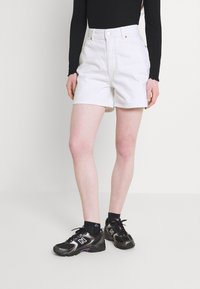 Valkeat denimmitrikoot, joissa on korkeavyötärö ja käännetty helma, yhdistettynä mustiin urheilukenkiin, joissa on violetit yksityiskohdat, sekä mustiin sukkiin.