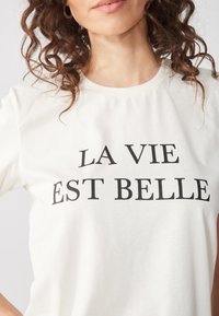 Camiseta de algodón blanca con el texto "LA VIE EST BELLE" en una fuente negra negrita en el pecho. Mangas cortas y escote redondo.