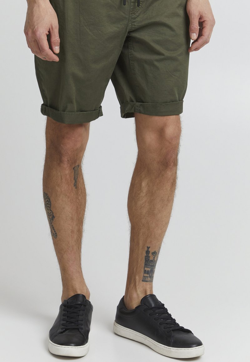 Shorts roulés vert olive en coton. Coupe slim avec poches, portés avec des baskets noires dotées de semelles blanches et de lacets texturés.