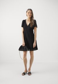 Vestido negro de encaje con mangas cortas abullonadas, escote en V y un corte holgado. Combinado con un bolso negro con cadena y sandalias negras.