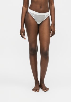 Kobieta stojąca boso, ubrana w białą, paskowaną bieliznę z wysokim stanem na jednolitym jasnym tle.