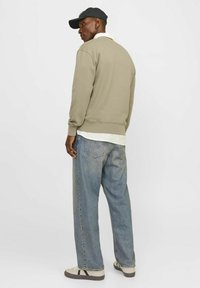 Sudadera caqui con puños acanalados, superpuesta a una camisa blanca, combinada con unos pantalones vaqueros azules holgados y zapatillas de perfil bajo. Una gorra negra completa el look.