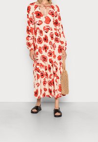 Femme portant une robe midi beige à imprimé floral rouge, à manches longues, décolleté en trou de serrure, tenant un sac tissé beige, vêtue de sandales noires.