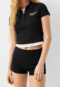 Femme portant un polo noir court avec le texte « 59th Street New York » et un short taille haute noir, montrant une partie d'un t-shirt blanc en dessous.