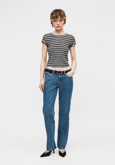 Γυναίκα με κοντά καστανά μαλλιά που φοράει ριγέ μαύρο και λευκό μπλουζάκι, μπλε τζιν με ζώνη και μαύρες μυτερές γόβες, στέκεται μπροστά σε λευκό φόντο.