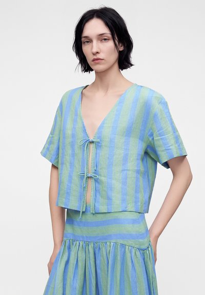Femme aux cheveux courts noirs portant un haut à manches courtes rayé verticalement bleu et vert, noué à l'avant, et une jupe assortie.