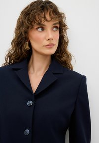 Granatowa marynarka szyta na miarę z klapami, trzema guzikami i gładką teksturą. Modelka nosi złote kolczyki w kształcie kółek, eksponując naturalne loki.