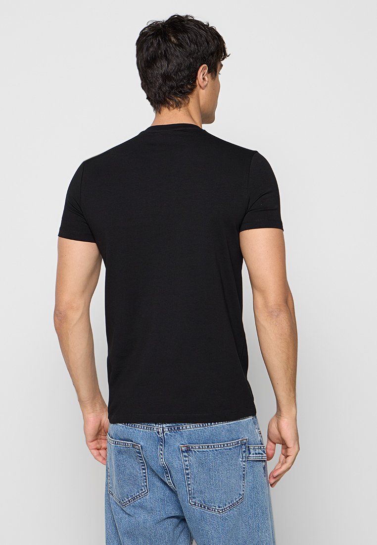 Hombre de espaldas vistiendo una camiseta negra lisa de manga corta y vaqueros de mezclilla azul claro sobre un fondo claro plain.