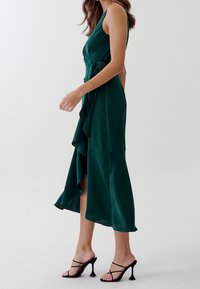 Kvinna som går i en mörkgrön ärmlös omslagklänning med volangdetaljer och svarta, remmade högklackade sandaler mot en vit bakgrund.