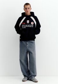 Ung person iført en sort Evangelion-hoodie, løse blå jeans og grå sneakers, stående mod en enkel hvid baggrund.