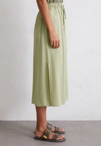 Light green midi skirt made of smooth fabric with a drawstring waistband and side slits. Paired with dark green slide sandals featuring gold accents.