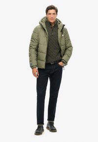 Oliven grøn pufferjakke med hætte, parret med en mørk striktrøje og mørke jeans. Skoene har mørke snørebåndsstøvler med lyse såler.