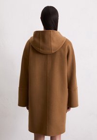 Manteau en laine marron avec capuche, silhouette oversize, épaules tombantes et manches larges. Texture lisse et lignes simples et épurées.