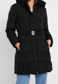 Abrigo tipo puffer negro con capucha, cintura ceñida, dos bolsillos laterales y cierre de botones a presión. Presenta un patrón acolchado horizontal y una textura suave.