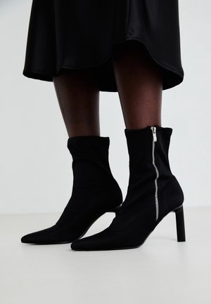 Bottes noires à bout pointu avec fermetures éclair latérales sur un modèle portant une jupe noire, sur un fond clair uni.