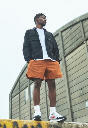 Chaqueta negra con múltiples bolsillos, camiseta blanca, pantalones cortos naranjas, zapatillas blancas y rojas, de pie contra una estructura de madera.