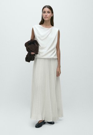 Kvinde iført ærmeløs hvid top og lang, plisseret hvid nederdel, der holder en mørkebrun jakke og står mod en ensfarvet lys baggrund.