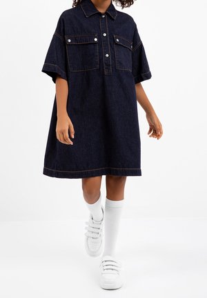 Jeansowa sukienka w ciemnym niebieskim kolorze z kołnierzykiem, krótkimi rękawami i dwiema przednimi kieszeniami. Stylizację dopełniają białe trampki i białe skarpetki do kolan.
