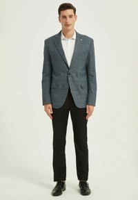 Homme portant un blazer à carreaux gris, une chemise blanche, un pantalon noir et des chaussures de ville noires, se tenant contre un fond clair uni.