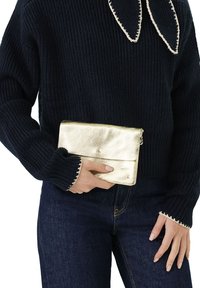 Gouden metallic clutch met een textuurfinish, bovenklep sluiting en stikdetails, in één hand gehouden. Draagt een donkere trui en spijkerbroek.
