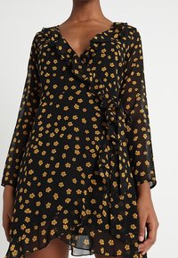 Černé zavinovací šaty s žlutým květinovým vzorem, volánkovým výstřihem a dlouhými rukávy. Průsvitná textura, asymetrický lem a postranní zavazování.