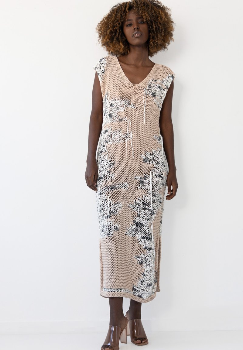 Beżowa sukienka maxi dzianinowa w czarno-białe kwiatowe wzory, z dekoltem w kształcie litery V, krótkimi rękawami typu cap oraz kieszeniami bocznymi. Przezroczyste buty na słupku.