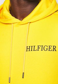 Felpa gialla con cappuccio e logo "HILFIGER" nero. Presenta coulisse con punte metalliche e tessuto morbido e strutturato.