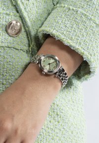 Ceas de mână cu brățară din metal argintiu, cu cadran verde și numere romane, purtat pe un încheietură, asortat cu o manșetă dintr-o jachetă de țesătură verde.