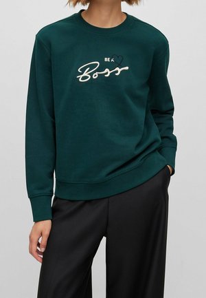 Persoon draagt een donkergroene sweatshirt met de tekst "Be a Boss" en een hartontwerp, gecombineerd met losse zwarte broek, handen ontspannen langs het lichaam.