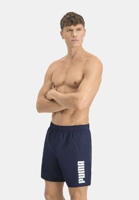 Los pantalones de baño de color navy, confeccionados en material de secado rápido, presentan una cinturilla elástica y un prominente logo blanco de "PUMA" en el lado izquierdo.