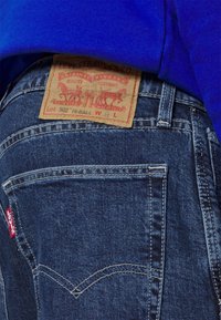 Jeans de mezclilla azul con una etiqueta roja de Levi's y un parche de cuero marrón claro con caballos, que exhibe la típica costura del bolsillo trasero.